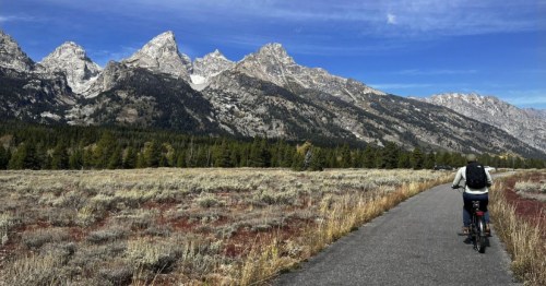 A cyclist rides along a path with mountains in the background and a clear blue sky above.