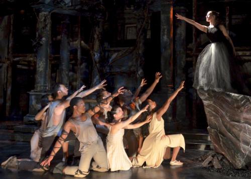 A dramatic stage scene with a woman in a black dress reaching out to a group of performers in various costumes below.