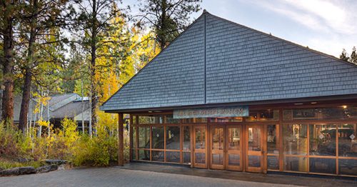 A modern building with a sloped roof, surrounded by trees and autumn foliage, showcasing large glass windows.