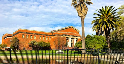 Historic brick building with columns, surrounded by palm trees and greenery, reflecting in a pond under a blue sky.