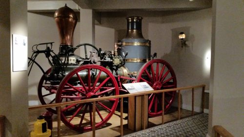 Historic steam engine on display, featuring large red wheels and a copper chimney, set in a museum environment.