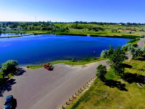 Aerial view of a serene lake surrounded by greenery and a parking area with a few cars under a clear blue sky.