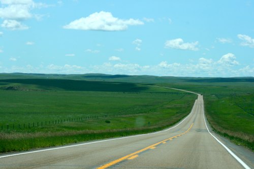 A long, winding road stretches through lush green fields under a bright blue sky with fluffy white clouds.