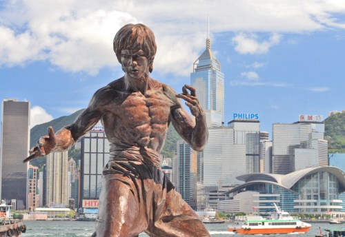 A bronze statue of a martial artist in front of a modern city skyline with skyscrapers and a harbor.