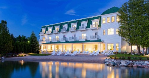 A charming hotel by a calm lake at dusk, featuring a green roof and illuminated windows, surrounded by trees.