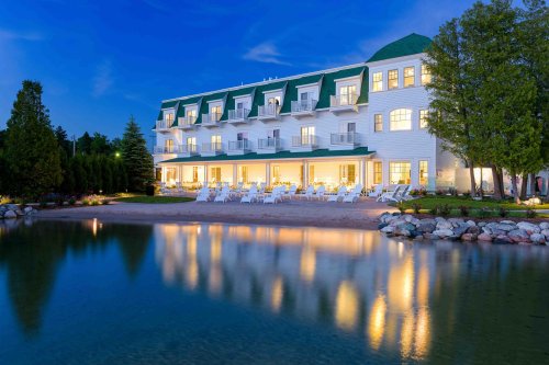 A charming hotel with green roofs, reflecting in a calm lake at dusk, surrounded by trees and outdoor seating.