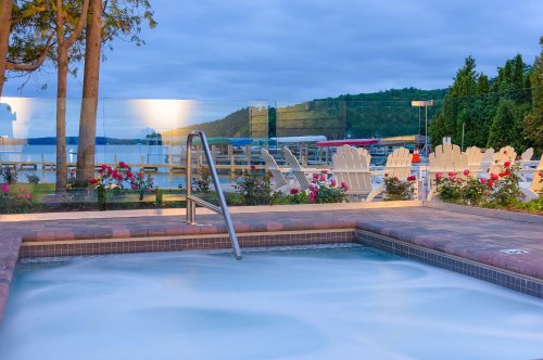 A serene hot tub with a view of a lake, surrounded by trees and flowers, under a cloudy sky.