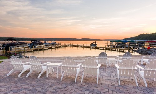 Adirondack chairs line a sandy beach, overlooking a calm lake at sunset with boats docked in the background.