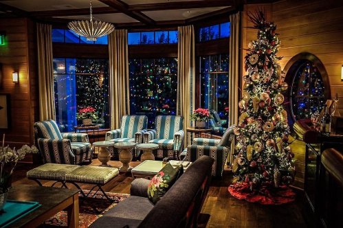 Cozy living room decorated for the holidays, featuring a Christmas tree and large windows with twinkling lights outside.