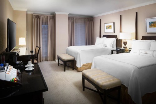 A hotel room featuring two beds, a desk, a TV, and elegant decor with soft lighting and neutral tones.