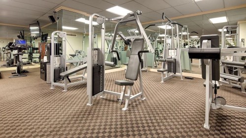 A spacious gym with various exercise machines and mirrors, featuring a clean, carpeted floor.