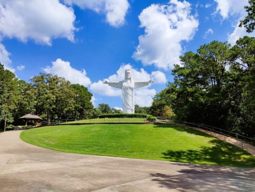 A large statue of Christ stands on a grassy hill under a blue sky with fluffy clouds, surrounded by trees.