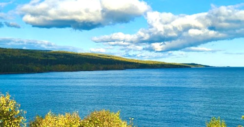 A serene view of a calm blue lake surrounded by green hills and fluffy clouds in a clear sky.
