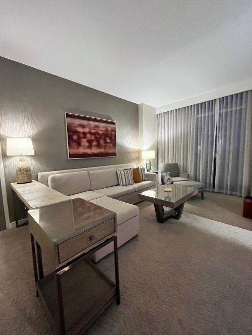 A modern hotel room with a light-colored sofa, glass coffee table, and large windows with sheer curtains.