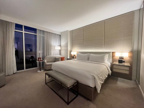 A modern hotel room featuring a king-sized bed, seating area, and large windows with sheer curtains.