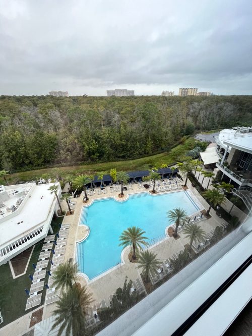 Aerial view of a hotel pool surrounded by palm trees, with a forested area in the background and cloudy skies above.