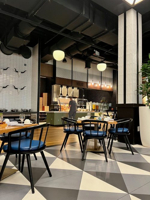 A modern restaurant interior with tables, a buffet area, and a staff member preparing food in the background.