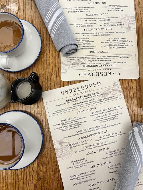 A table setting with two menus, coffee cups, a creamer, and gray napkins on a wooden surface.