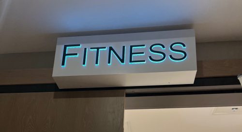 Sign for a fitness room with illuminated "FITNESS" above the door and a plant visible inside.