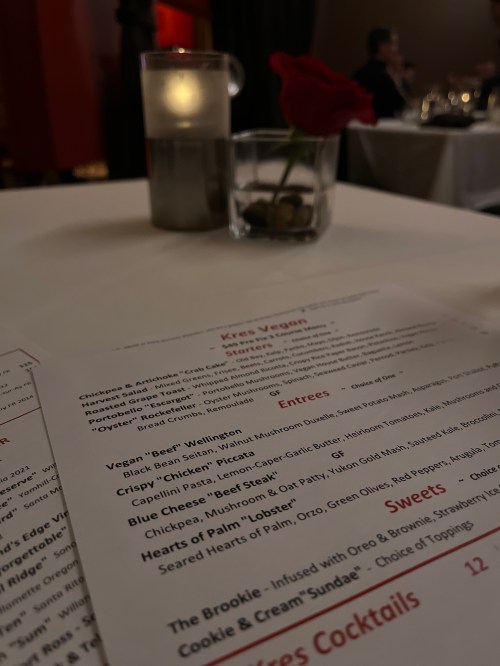 A table setting with a menu, candle, and a rose in a restaurant, featuring vegan dishes and desserts.