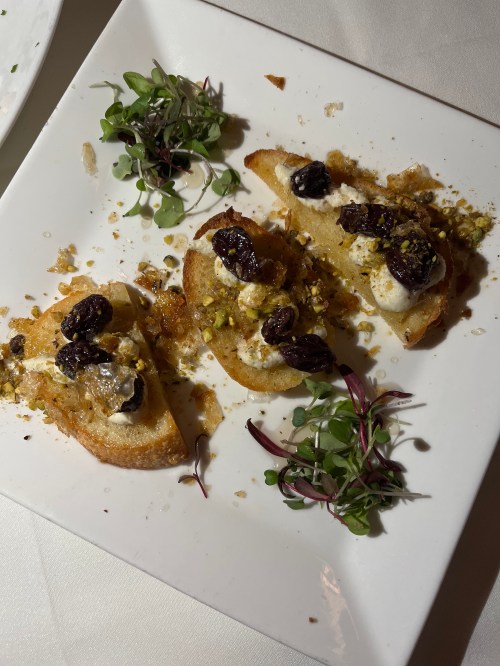 Three slices of toasted bread topped with cream, raisins, pistachios, and microgreens on a white plate.
