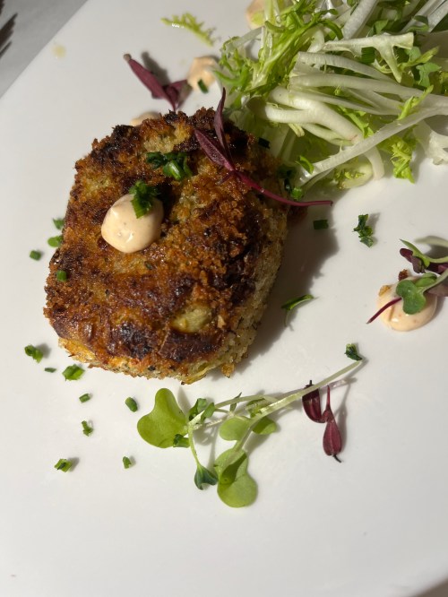 A golden-brown croquette topped with sauce, garnished with microgreens and a side of fresh salad.