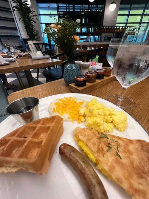 A plate with a waffle, scrambled eggs, sausage, and cheese, with a glass of water and jars of jam in the background.