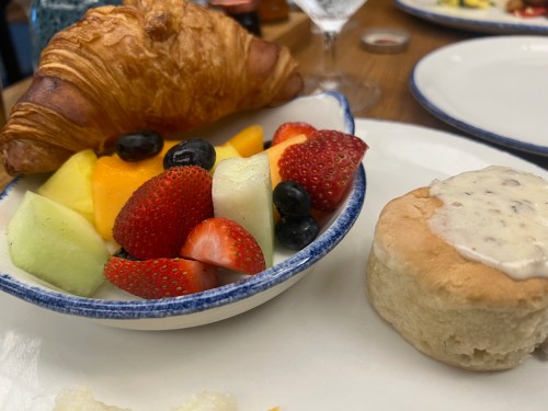 A plate with a croissant, a bowl of mixed fruit, and a biscuit topped with cream.