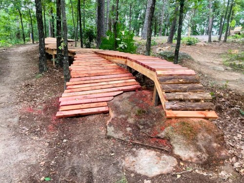 A wooden bridge made of planks curves over a rocky area, surrounded by trees and natural terrain.