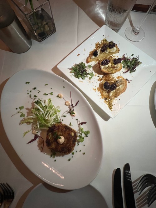 A plate of crab cakes with salad and a dish of crostini topped with blueberries and microgreens on a dining table.