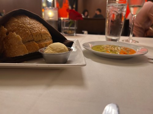 A plate with a bread roll and butter beside a bowl of soup on a table set for dining.