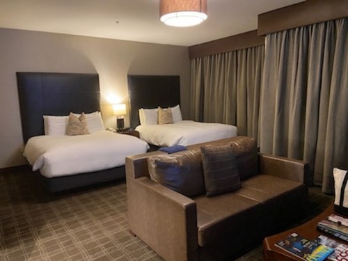 A hotel room featuring two beds, a sofa, and curtains, with warm lighting and a cozy atmosphere.