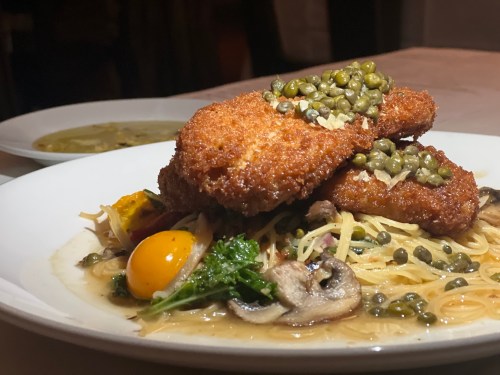 A plate of pasta topped with two breaded chicken cutlets, capers, and a side of soup.