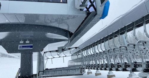 A ski lift station with empty chairs, surrounded by falling snow in a winter landscape.