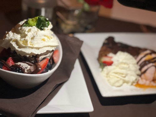 A dessert bowl with whipped cream, strawberries, and chocolate, alongside a slice of cake with whipped cream on a plate.