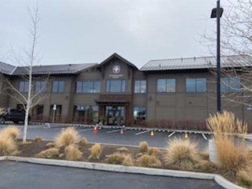 A modern building with large windows and a clock, surrounded by sparse landscaping and a parking lot.