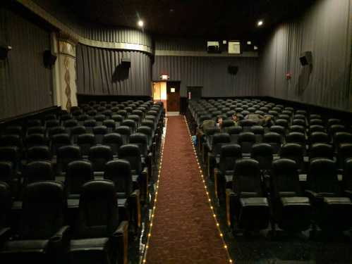 An empty movie theater with rows of black seats and a dimly lit aisle leading to the exit.