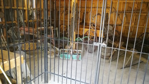 A rustic storage shed filled with old tools and equipment, viewed through a black metal gate.