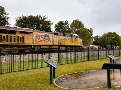 A train with an American flag design passes by a park with informational plaques and green grass.