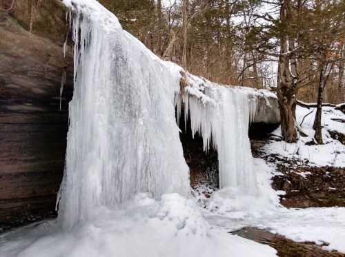 A frozen waterfall with icicles, surrounded by snow-covered rocks and trees in a winter landscape.
