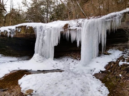 Frozen waterfall with icicles, surrounded by snow-covered rocks and trees in a winter landscape.