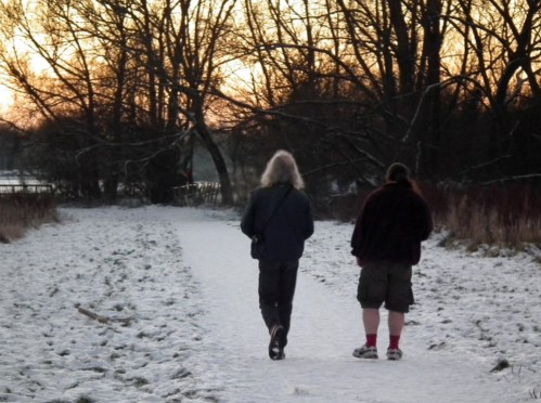 Two people walking on a snowy path at sunset, surrounded by trees and a serene winter landscape.