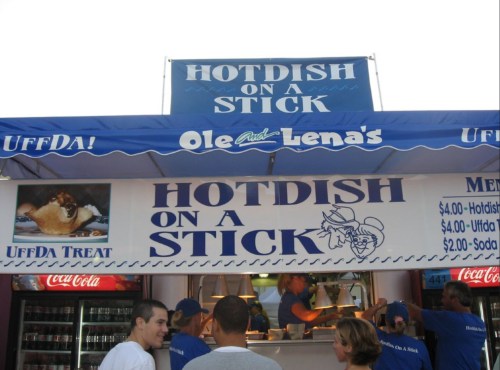 A food stand sign reads "Hotdish on a Stick" with menu prices and a cartoon character, surrounded by customers.