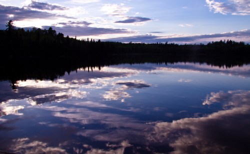 A serene lake reflecting clouds and trees under a blue sky at sunset.