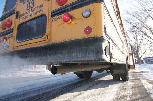 A yellow school bus on a snowy road, emitting exhaust smoke from its rear.