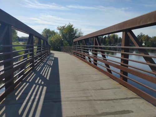 A metal bridge with wooden railings spans a calm river, surrounded by trees and a clear blue sky.