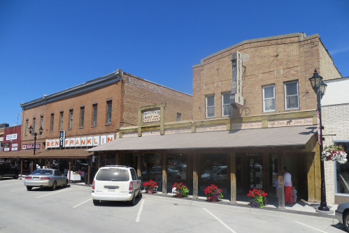 Historic buildings line a sunny street, featuring a store with "Ben Franklin" signage and colorful flower planters.