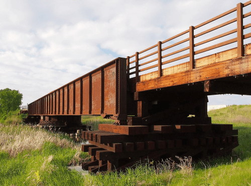 Rusty train bridge supported by wooden beams, surrounded by tall grass and a cloudy sky.