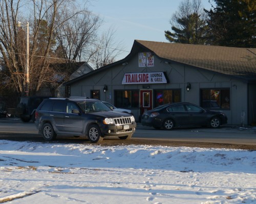 A gray building with a sign reading "Trailside Lounge & Grill," surrounded by snow and parked cars.