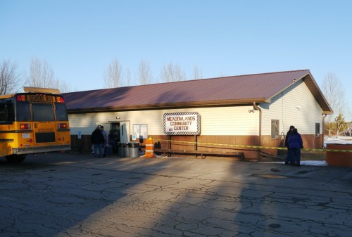 A community center building with a sign, a school bus parked nearby, and people entering the center on a clear day.